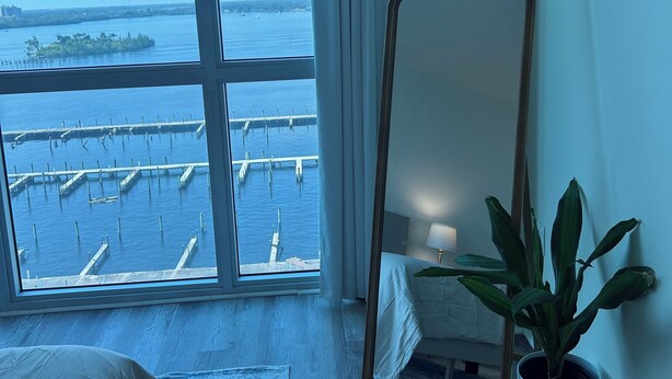 A hotel room with a large window overlooking a marina, a bed, a mirror, and a potted plant. - Tan Haven-A Chic & Cozy Modern Getaway (Fort Myers)