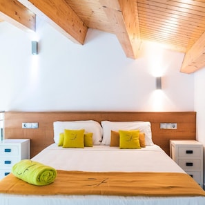 Une chambre d’hôtel avec un plafond en bois, un lit avec une literie blanche et des oreillers jaunes, une serviette enroulée et une table de chevet avec un tiroir.