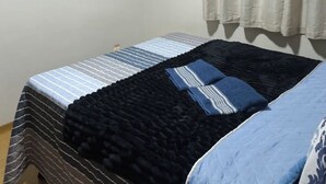 Un dormitorio con cama, manta azul y negra, aire acondicionado blanco y cortinas blancas.