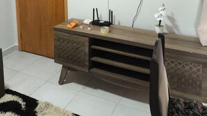 Un salón con puerta de madera, televisor de pantalla plana montado en la pared, una pequeña estantería con una planta en maceta y un mueble de madera con dispositivos electrónicos.