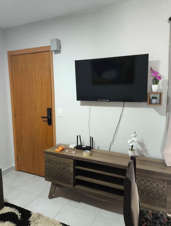 A living room with a wooden door, a flat-screen TV mounted on the wall, a small shelf with a potted plant, and a wooden entertainment unit with electronic devices.