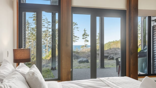 A bedroom with a bed, white pillows, and a view of the ocean through large glass doors. - Pacific Seas Suite-Wild Pacific Trail, Ocean View! (Ucluelet)