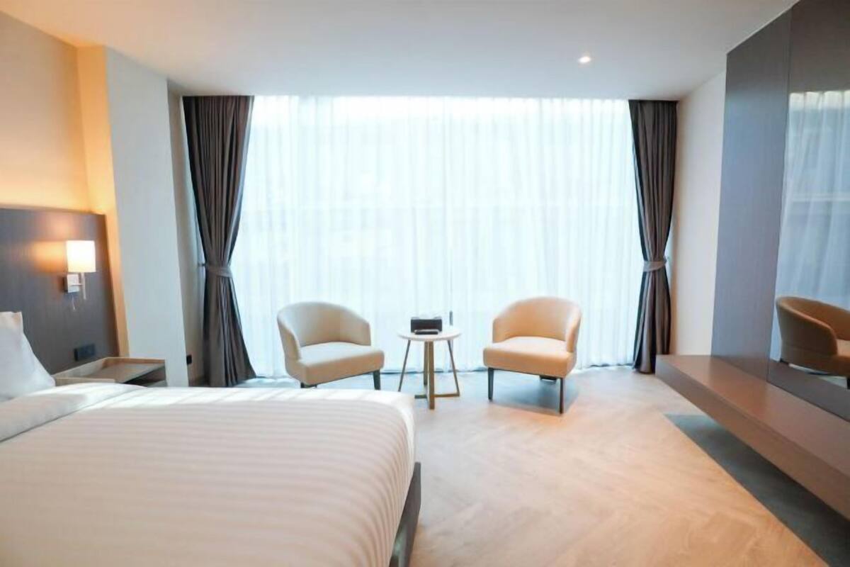 Una habitación de hotel con una cama grande, dos sillones, una mesita y un ventanal con cortinas.