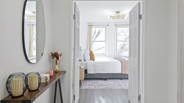 Un pasillo que conduce a un dormitorio con una cama, una mesita de noche con objetos decorativos y un espejo grande en la pared.