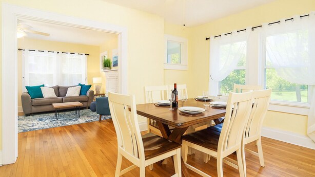 Un comedor con una mesa de madera dispuesta para cuatro, sillas blancas y un sofá al fondo.