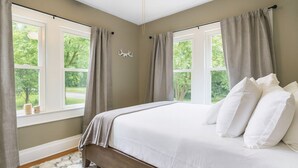 Un dormitorio con una cama grande, dos ventanas con cortinas, un ventilador de techo y vistas a zonas verdes al exterior.