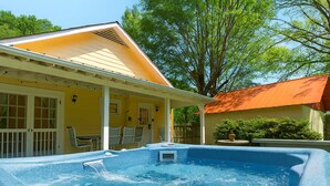 Una piscina con un jacuzzi, un porche cubierto con sillas, y una casa amarilla con techo rojo.