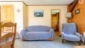 Et hyggeligt rum med en sofa med træramme, en lænestol og et spisebord med stole. Et maleri hænger på væggen, og en trædør fører til et andet rum.