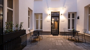Une cour avec une clôture en métal noir, deux tables rondes avec des chaises et une porte surmontée d’une lampe.