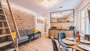 Un salon moderne avec un mur en briques, un escalier en bois, un canapé gris et un coin repas avec une table en bois mise pour deux personnes.