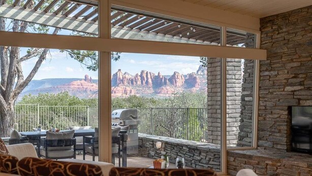 A spacious living area with a stone fireplace, large windows offering a view of red rock formations, and outdoor seating with a grill. - New Listing Vast Red Rock Views 2 Master Suites - Panorama Landing (Sedona)
