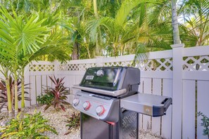 A barbecue grill with a digital display and control knobs, set against a white fence and surrounded by tropical plants.