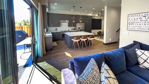 Une cuisine moderne avec des meubles de couleur sombre et un grand îlot central, un coin repas avec un canapé bleu et des coussins à motifs, et une vue sur une cour arrière équipée d’un barbecue.