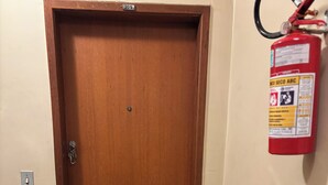 A wooden door with a keyhole and a lock, a fire extinguisher mounted on the wall, a potted plant, and a grey rug on the floor. - 2-Bedroom Apartment Near Ouro Minas Hotel (Belo Horizonte)