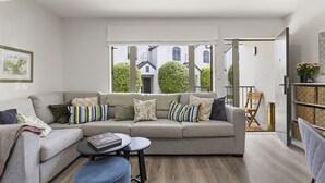 Una sala moderna con un sofá gris, varios cojines, una mesita redonda y un reposapiés azul. Un ventanal grande ofrece vistas a una zona residencial.