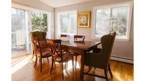 A dining area with a wooden table and chairs, large windows, and a painting on the wall. - Manchester by the Sea l Fireplace l King Bed (Manchester-by-the-Sea)