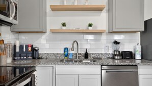 Una cocina moderna con armarios blancos, encimeras de granito, lavavajillas de acero inoxidable, horno integrado, microondas y cafetera. También hay dos pequeñas macetas con plantas en la encimera.