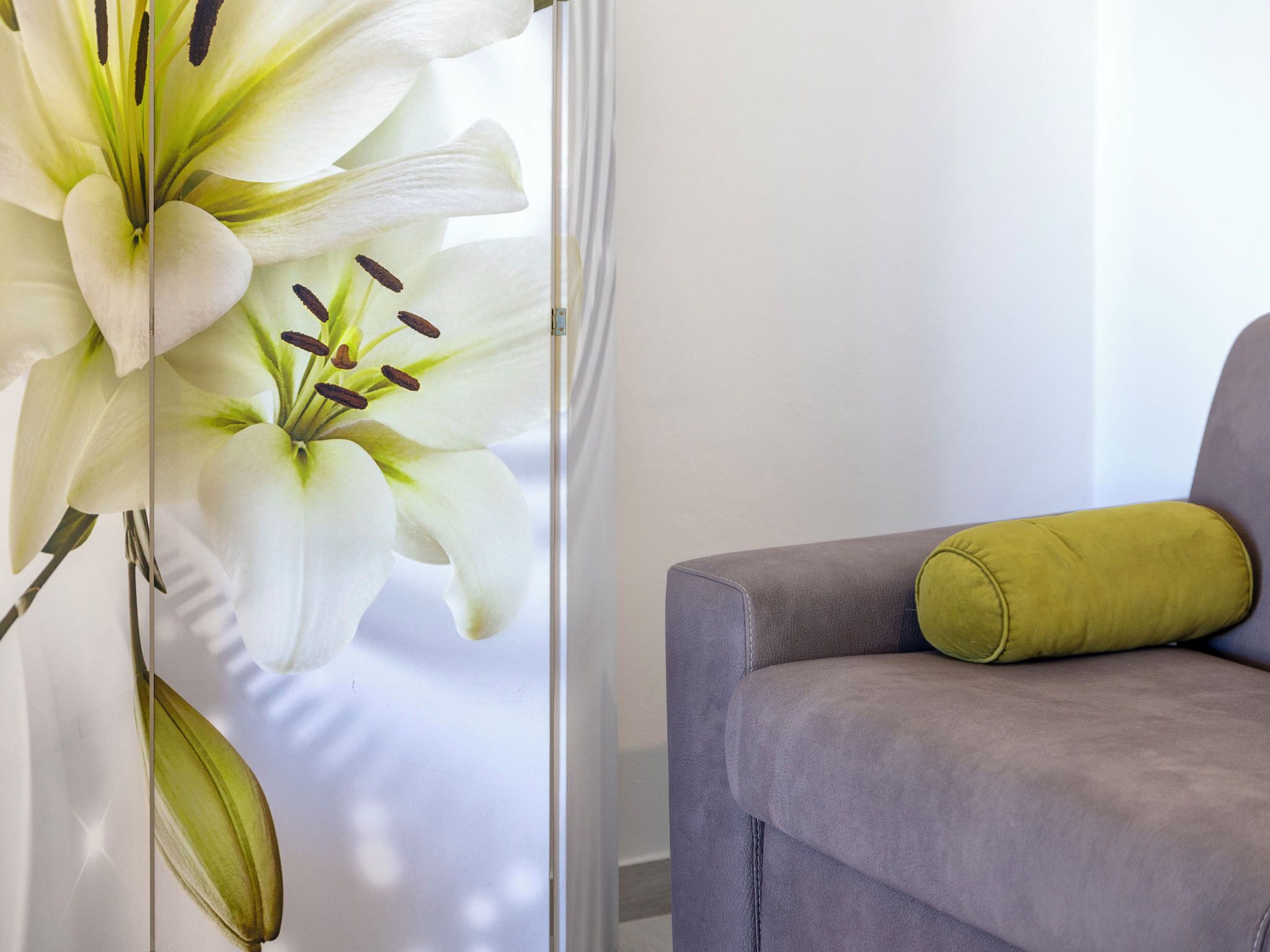 Una habitación con un sofá gris y un cojín verde, un arreglo floral blanco grande y una ventana con cortinas traslúcidas.