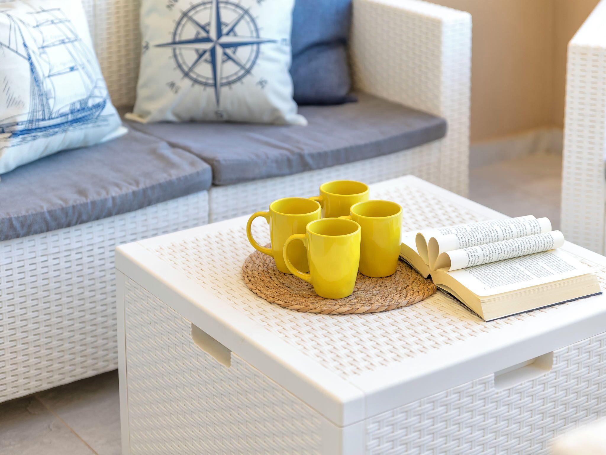 Ein weißes Rattan-Sofa mit einem Kissen mit Kompassmuster, eine gewebte Unterlage mit gelben Tassen und ein offenes Buch auf einer weißen Kiste.