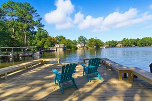 Une jetée en bois avec deux fauteuils Adirondack verts et un banc, d’où l’on peut admirer un lac paisible, avec des bateaux et des maisons en arrière-plan.