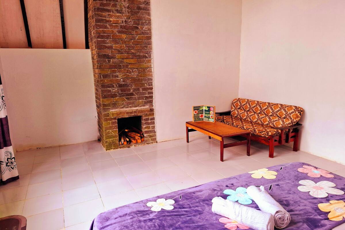 A room with a brick fireplace, a sofa, a small wooden table, and a purple floral bedspread.