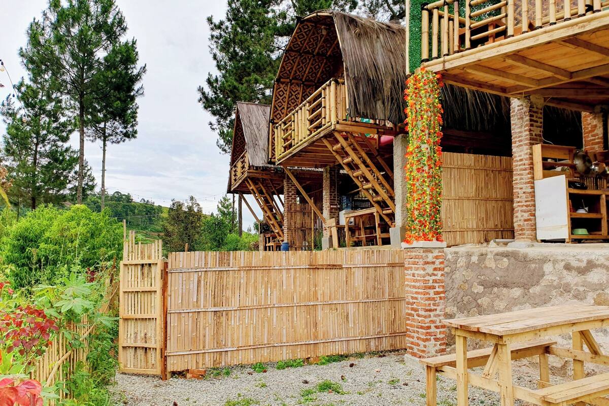 A rustic wooden house with a thatched-roof structure, a wooden fence, and a gravel yard.
