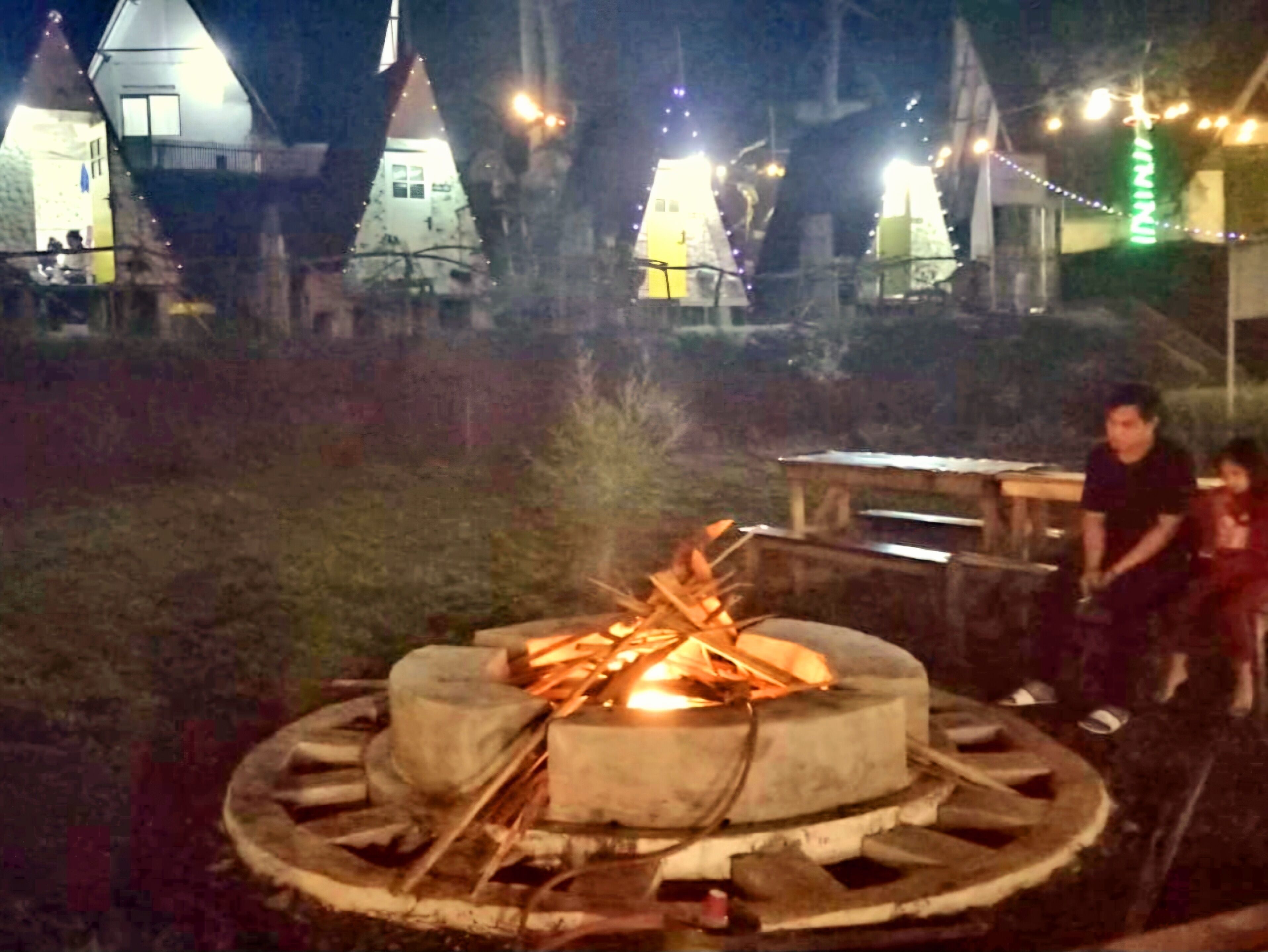 A campfire in a circular stone fire pit surrounded by logs, with a wooden bench and a table nearby.