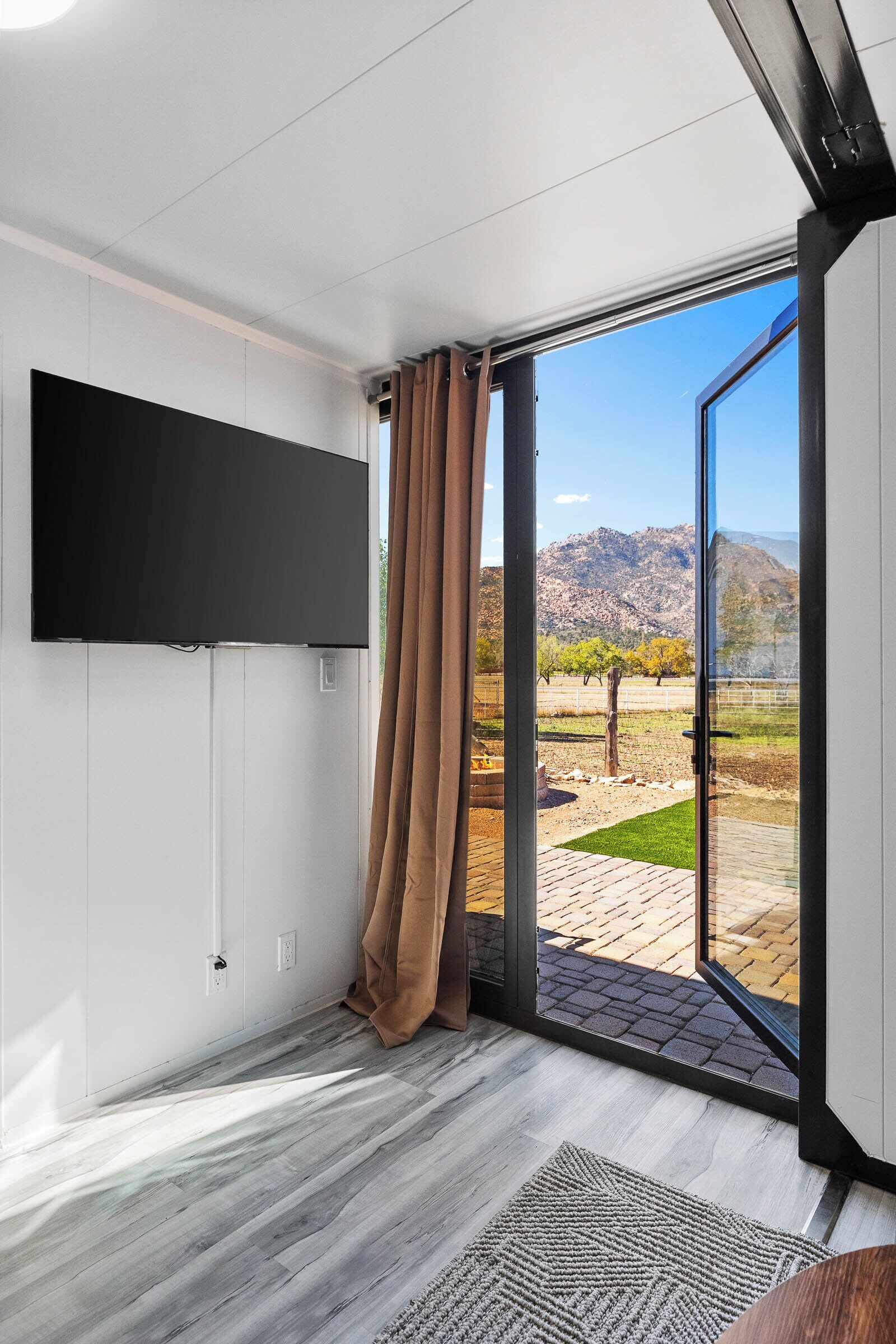 Una habitación moderna con televisor de pantalla plana en la pared, puerta corredera de vidrio hacia un espacio exterior con vistas a montañas y árboles.