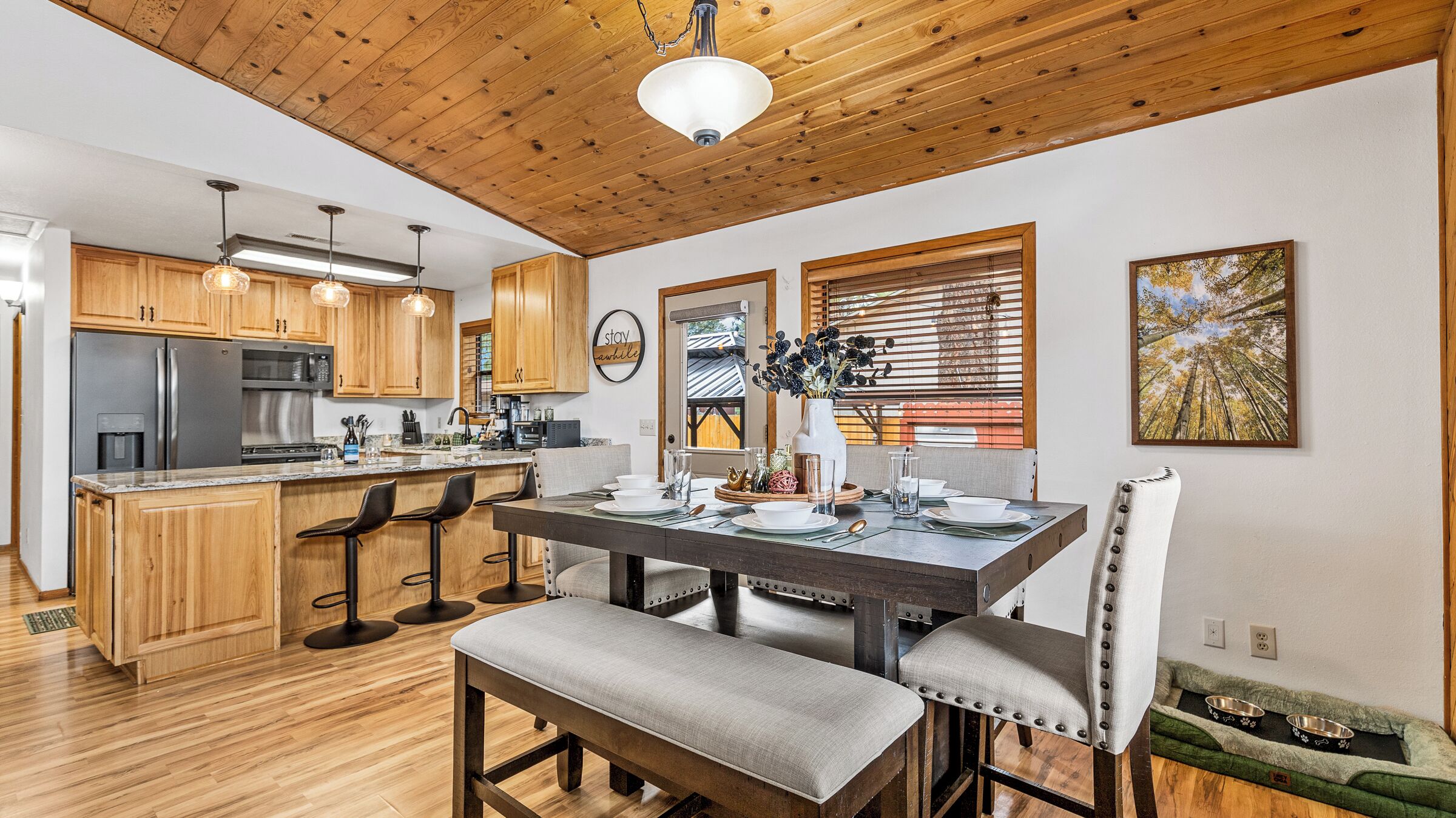 A modern kitchen with wooden cabinets, a central island with bar stools, a dining table set for four, and a pet bed.