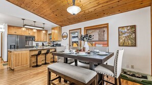 A modern kitchen with wooden cabinets, a central island with bar stools, a dining table set for four, and a pet bed.