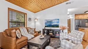 A living room with a brown leather sofa, a plaid armchair, a wooden coffee table, and a TV on the wall.