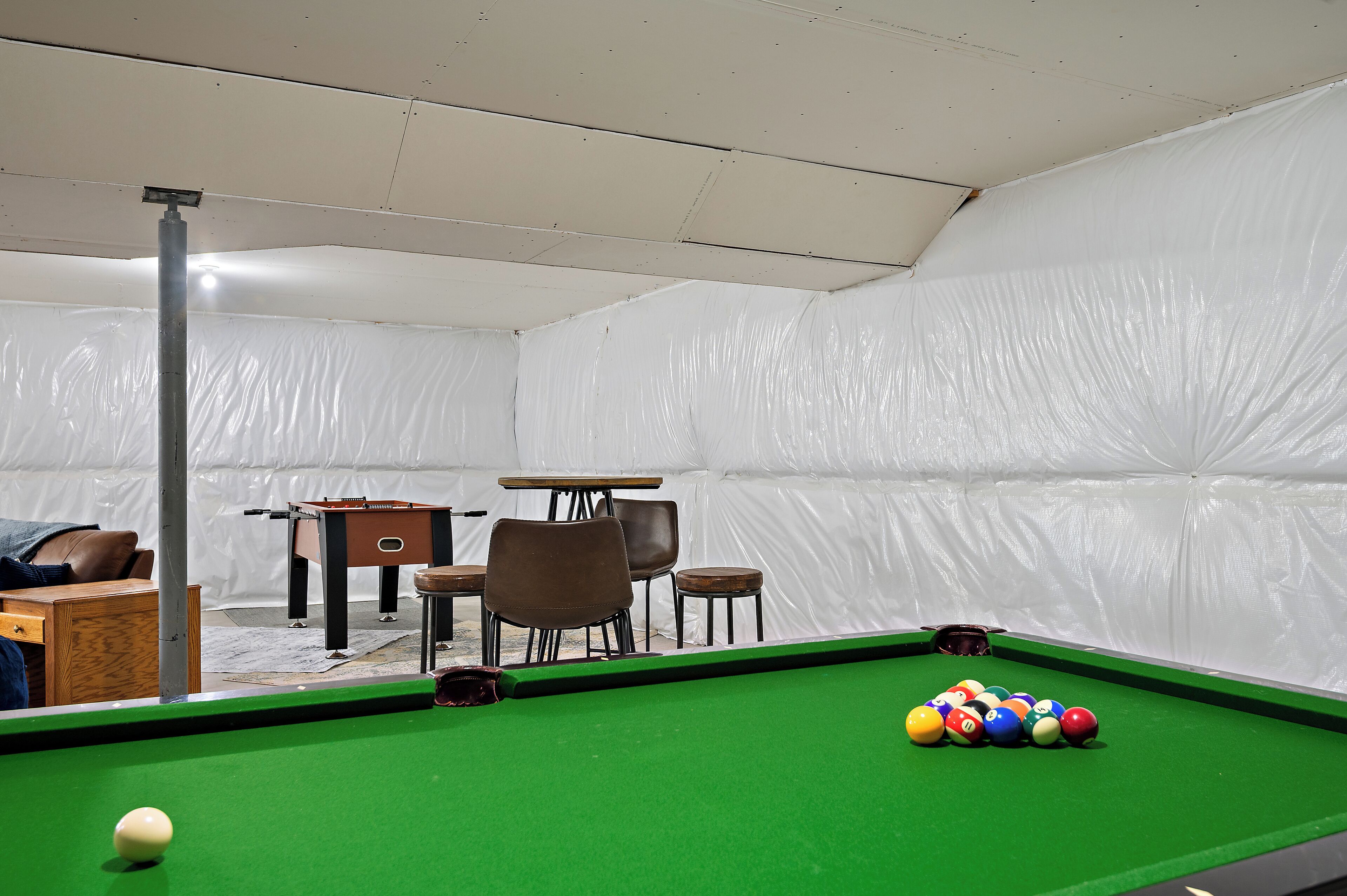 A pool table with balls, a pool cue, and a table with chairs in a room with white walls.