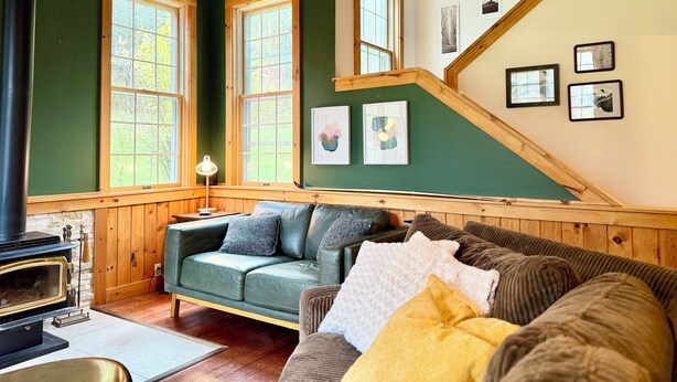Un salon chaleureux avec un mur vert, un plafond en bois et une cheminée. On y trouve deux fenêtres, un escalier et des tableaux encadrés accrochés au mur.