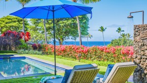 A poolside area with a blue umbrella, two wicker lounge chairs, a stone wall, and a view of the ocean.