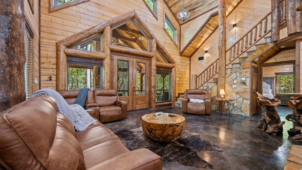 A spacious log cabin interior with leather sofas, a central coffee table, and a stone fireplace.