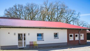 A single-story building with a red roof, a small porch with a bench, and a dirt ground.