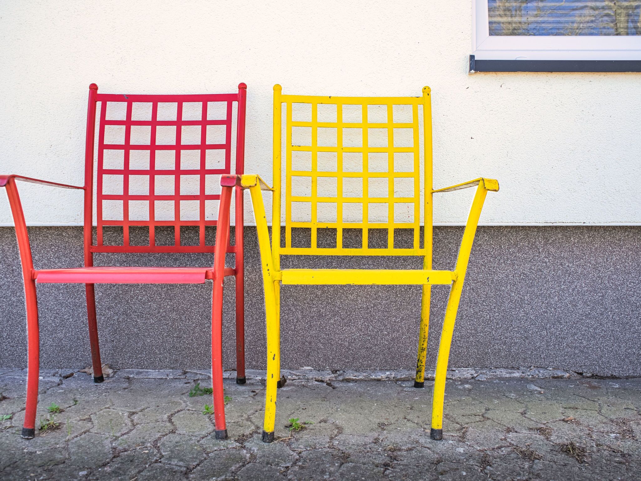 Deux chaises, une rouge et une jaune, sont placées sur un pavé, adossées à un mur blanc.