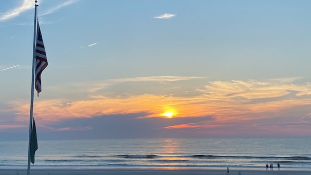 Ein Sonnenuntergang über dem Meer mit einer Flagge auf einem Mast, einer Promenade und Menschen, die am Strand entlanggehen.