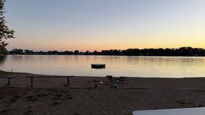 Vista de un lago con muelle, mesas de picnic y una playa arenosa.