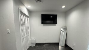 A small, modern room with a flat-screen TV mounted on the wall, a large mirror, and a trash bin. - Clewiston Comfort #4 – Stylish 1BR Hideaway (Clewiston)