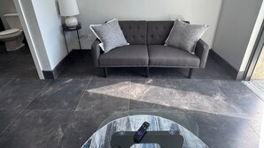 A modern living room with a grey sofa, two grey cushions, a side table with a lamp, and a remote control on a glass table. - Clewiston Comfort #4 – Stylish 1BR Hideaway (Clewiston)