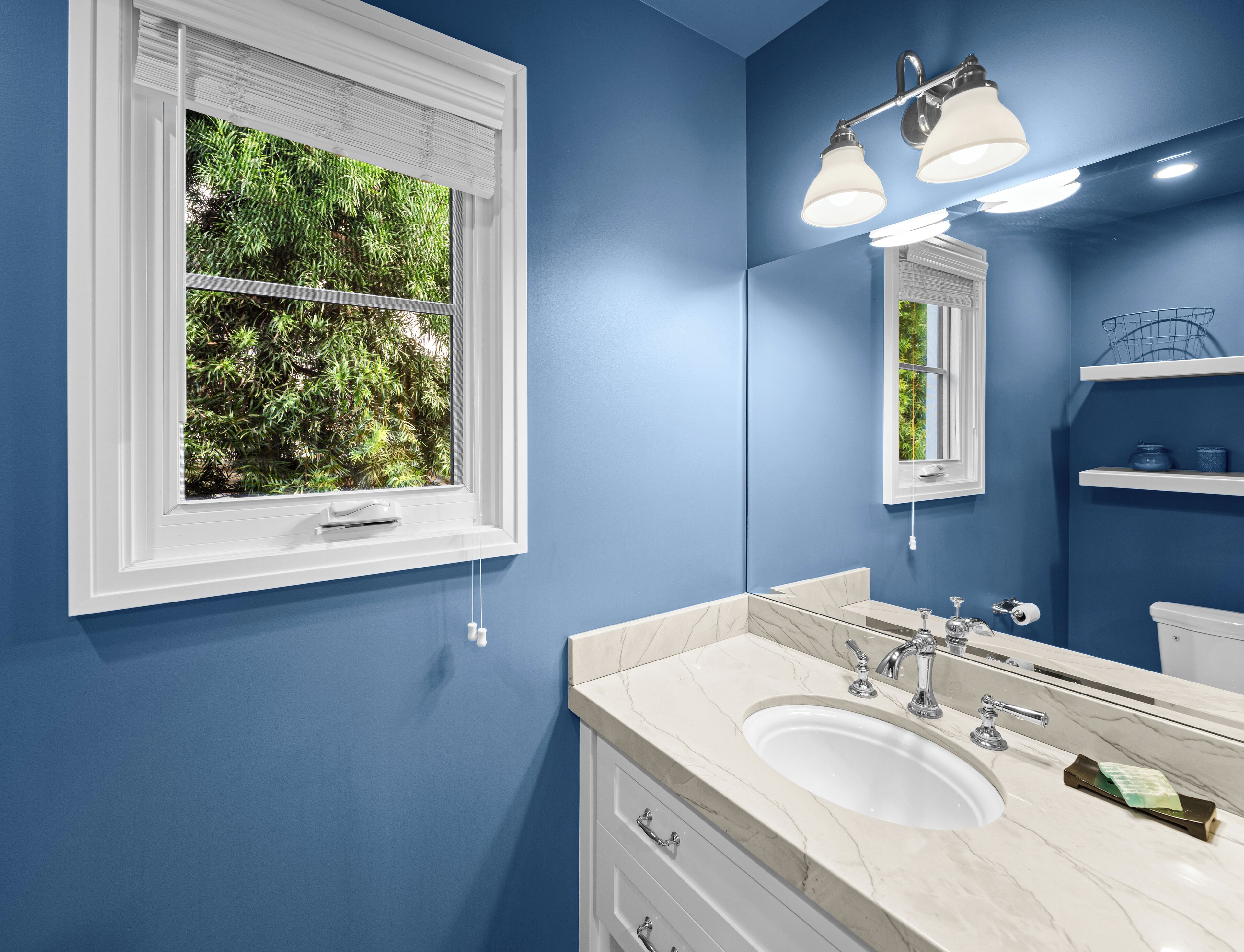 Un bagno con una parete blu, un mobile lavabo bianco con lavandino, uno specchio e una finestra con persiane.