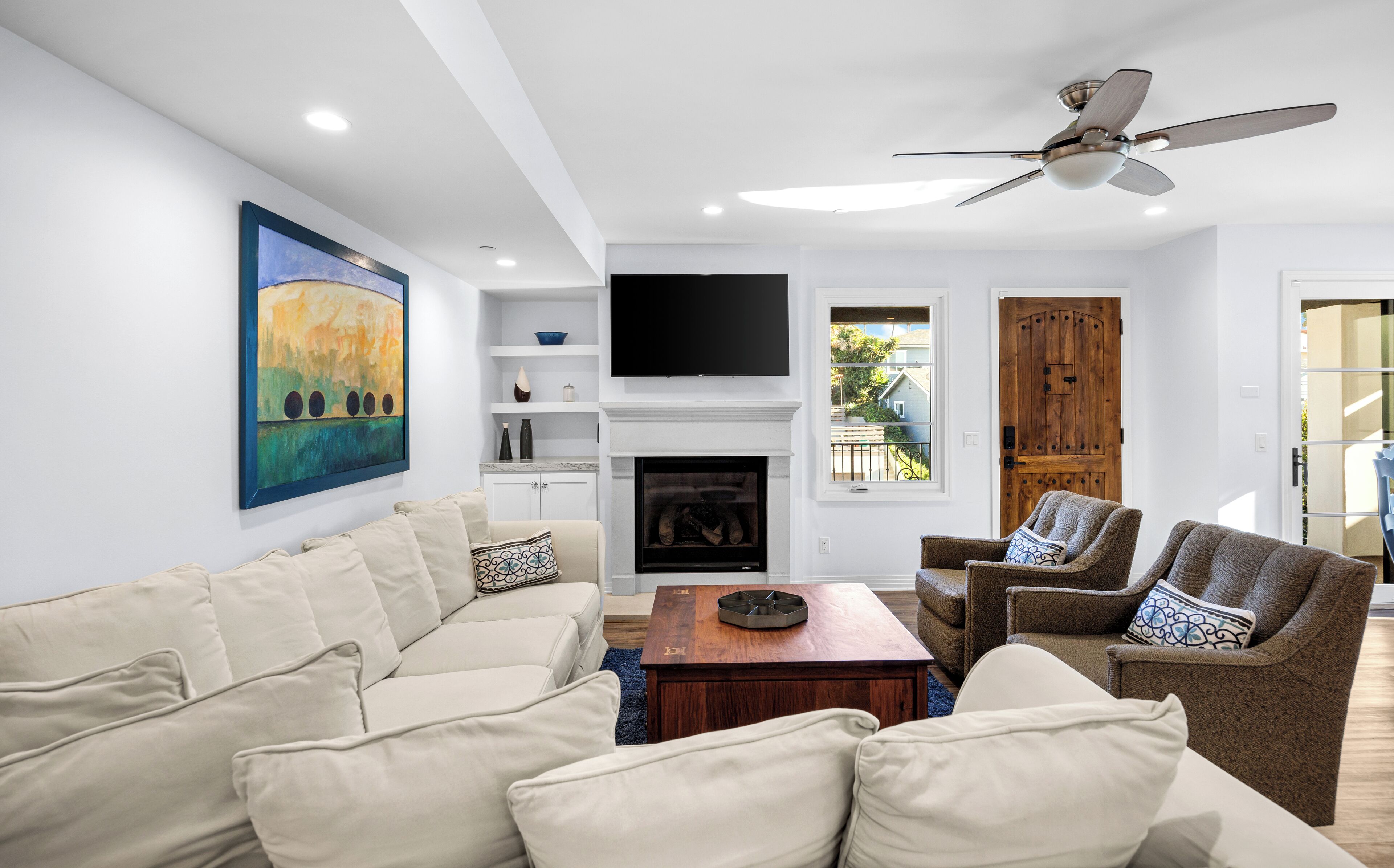 A modern living room with a beige sofa, a brown armchair, a wooden coffee table, a fireplace, a flat-screen TV, and a ceiling fan.