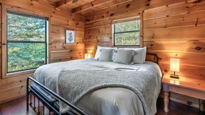 A wooden cabin bedroom with a bed, two windows, a ceiling fan, and a nightstand with a lamp. - Dog Friendly Cabin - Close to Gatlinburg - Fire pit - Outdoor Movie Projector (Sevierville)