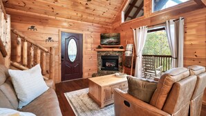 A cozy cabin interior with a wooden staircase, a fireplace, a sofa, and a window with curtains. - Dog Friendly Cabin - Close to Gatlinburg - Fire pit - Outdoor Movie Projector (Sevierville)