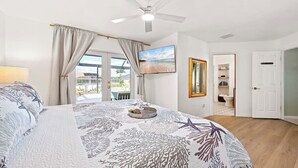 A bedroom with a bed, a ceiling fan, a window with curtains, a painting, a mirror, and a door leading to a bathroom. - Casa Azul - 3 Bedroom + Den 2 Bath Coastal Flair (Cape Coral)