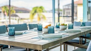 A dining table set with plates, glasses, and a bowl of food, with a view of the sea and palm trees in the background. - Casa Azul - 3 Bedroom + Den 2 Bath Coastal Flair (Cape Coral)