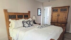 A neatly made bed with white bedding, a wooden headboard, and decorative pillows. There is a wooden wardrobe with intricate carvings to the right of the bed.