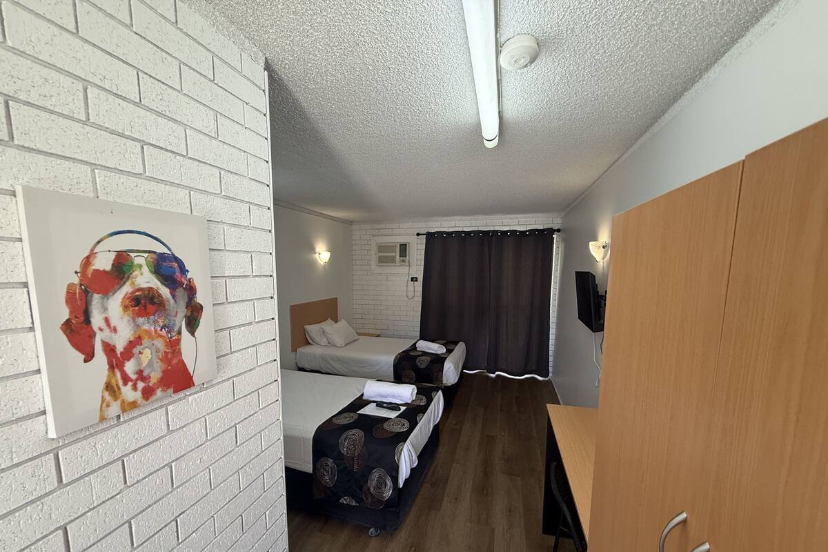 Una habitación de hotel con pared de ladrillo blanco, un cuadro de un perro, dos camas, un escritorio, un armario de madera y una cortina.