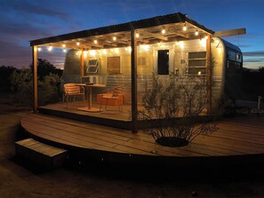 Une petite terrasse en bois, bien éclairée, avec une table et des chaises, entourée d’une structure métallique décorée de guirlandes lumineuses.